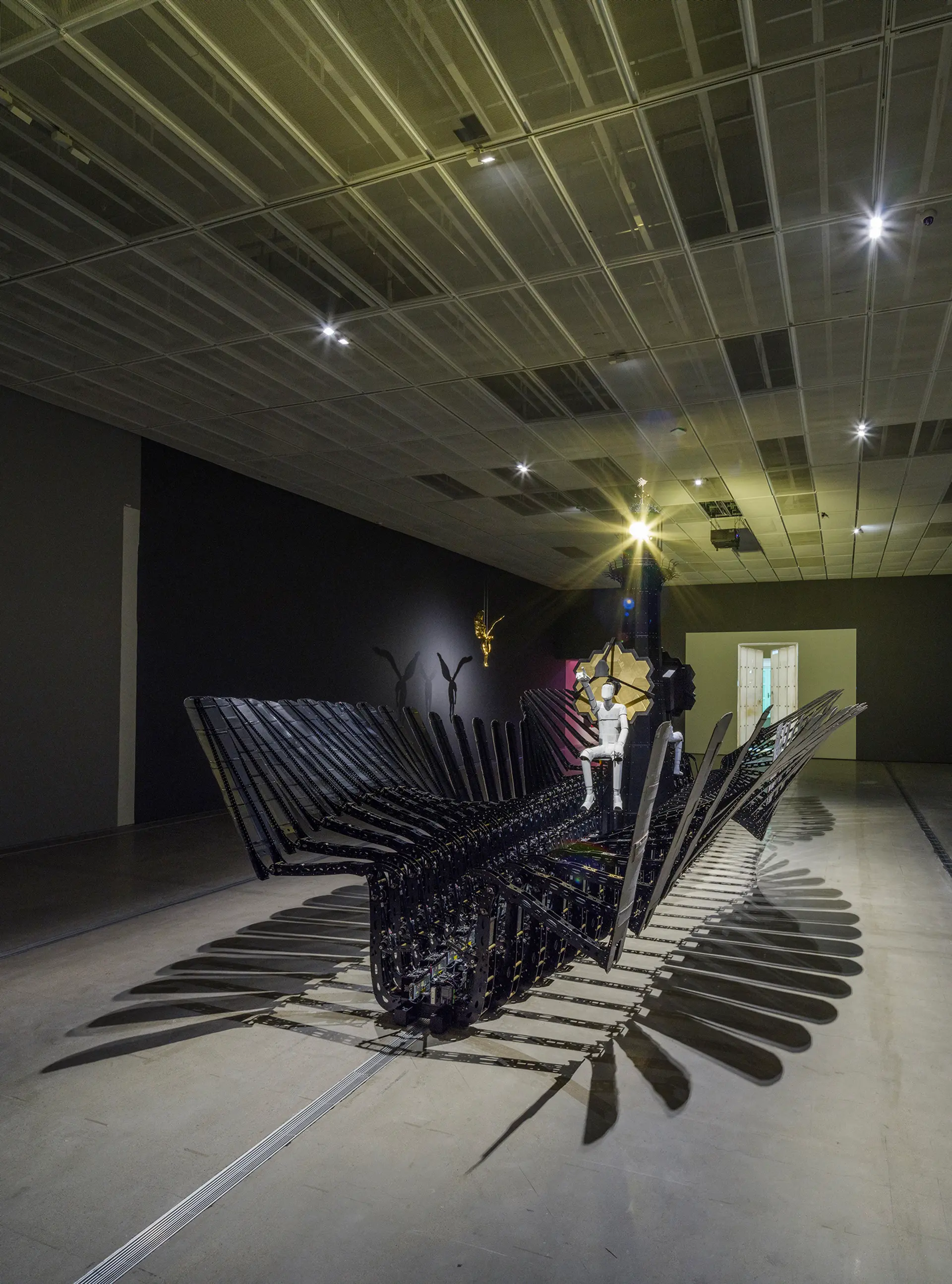 An industrial-style installation resembling an ark with a human-like statue seated in it. Overhead, a golden angel hangs from the ceiling.