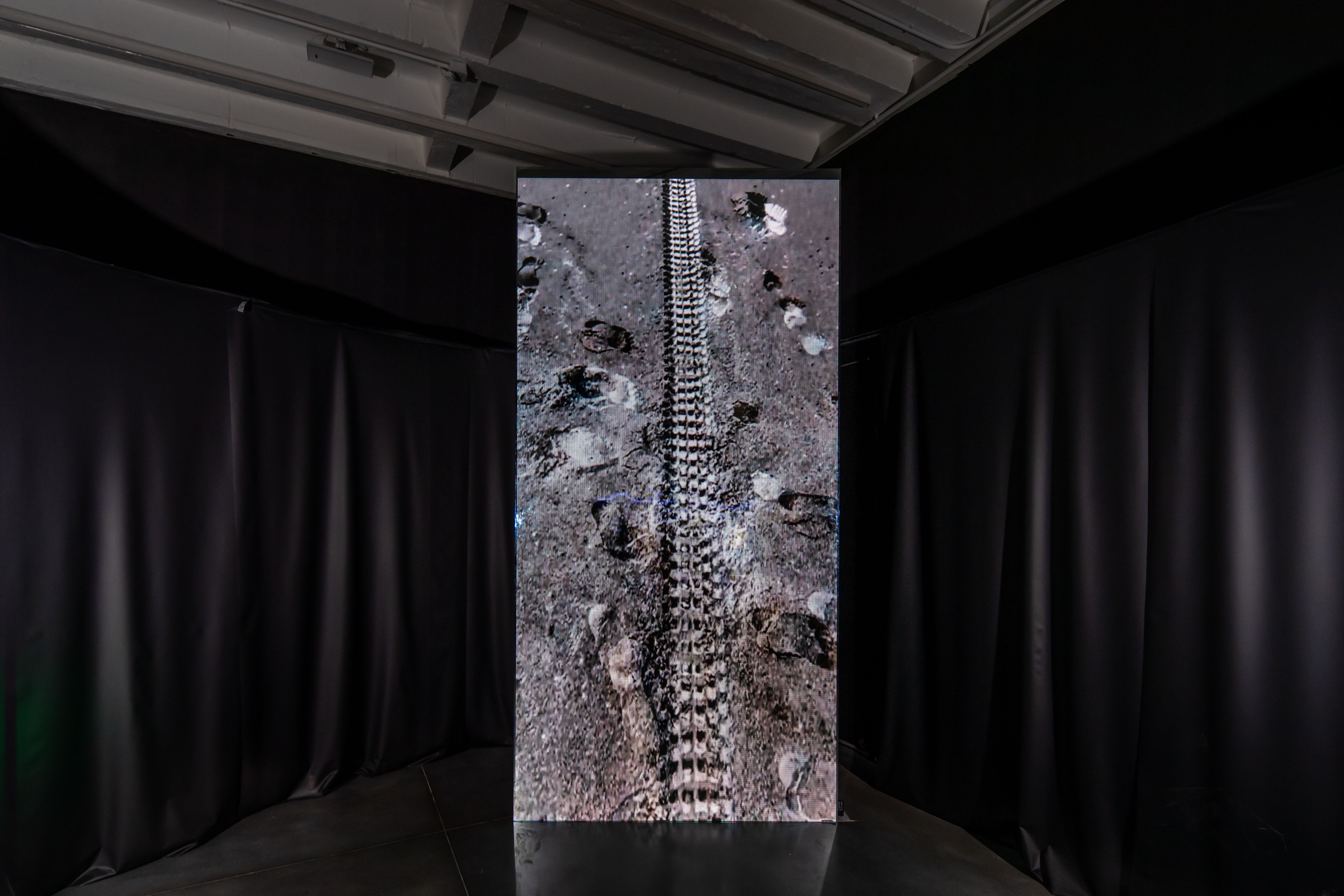 An art installation featuring a dark floor imprinted with footprints and a tire track, creating an aesthetic reminiscent o...