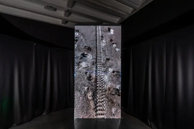 An art installation featuring a dark floor imprinted with footprints and a tire track, creating an aesthetic reminiscent o...