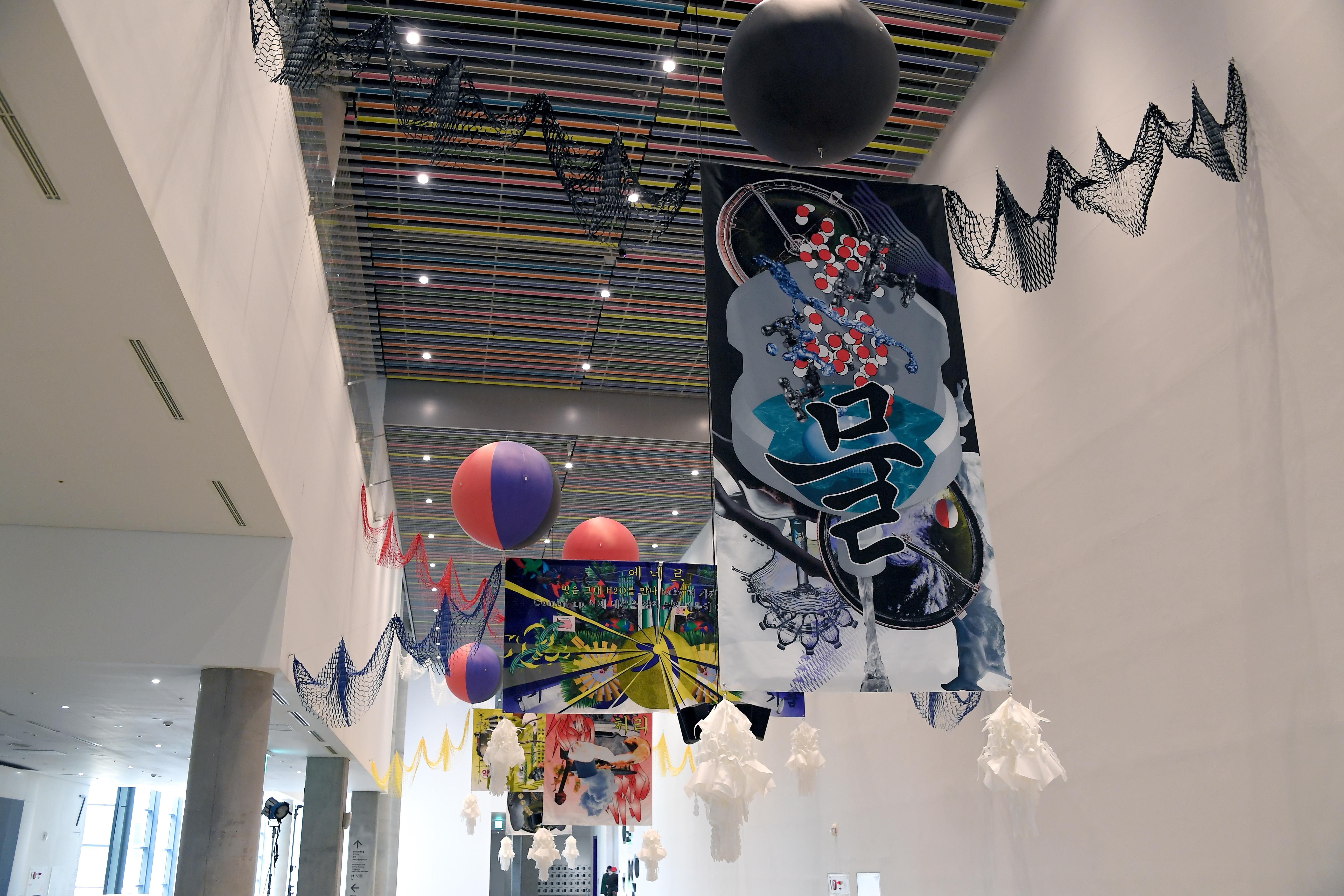 Numerous colorful, geometric art installations suspend from the ceiling in an exhibition space. Two bright orbs hover amid...