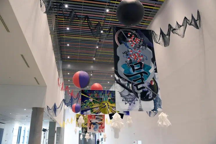 Numerous colorful, geometric art installations suspend from the ceiling in an exhibition space. Two bright orbs hover amid...