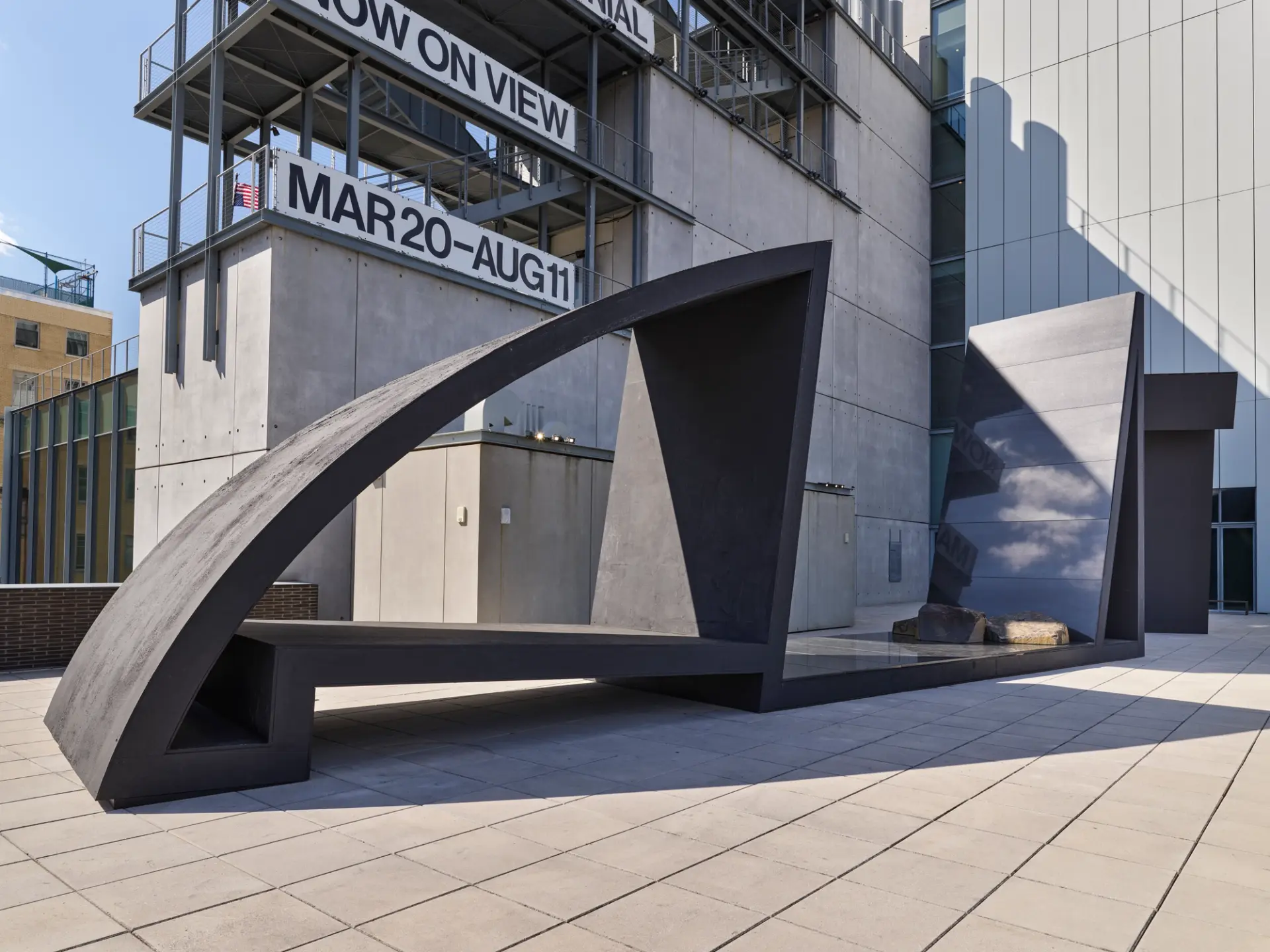 A large, black abstract outdoor sculpture with curved and angled sections, located on a rooftop terrace with industrial surroundings.