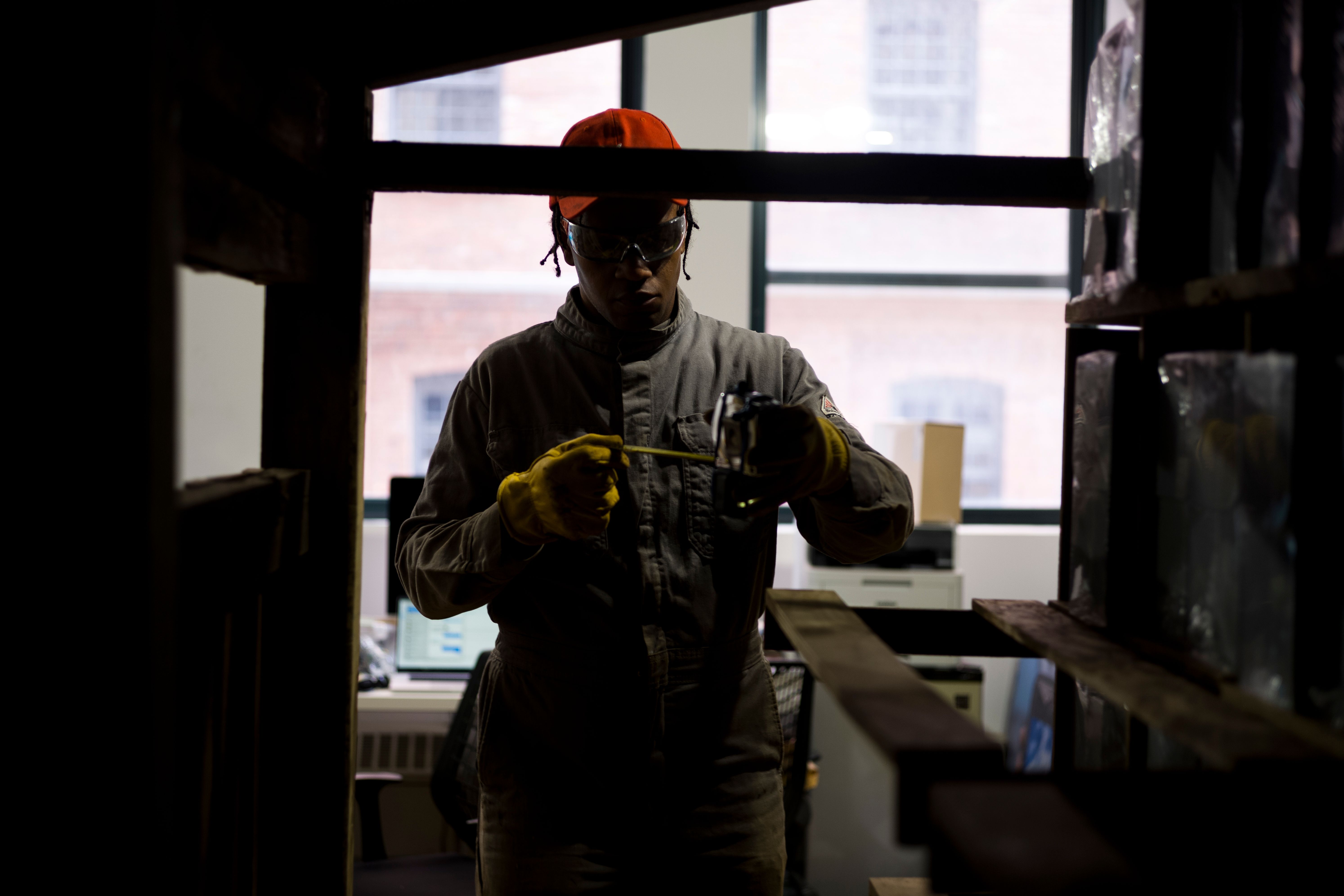 A worker in a protective suit and gloves measures something with a tool, in a dimly lit workshop.