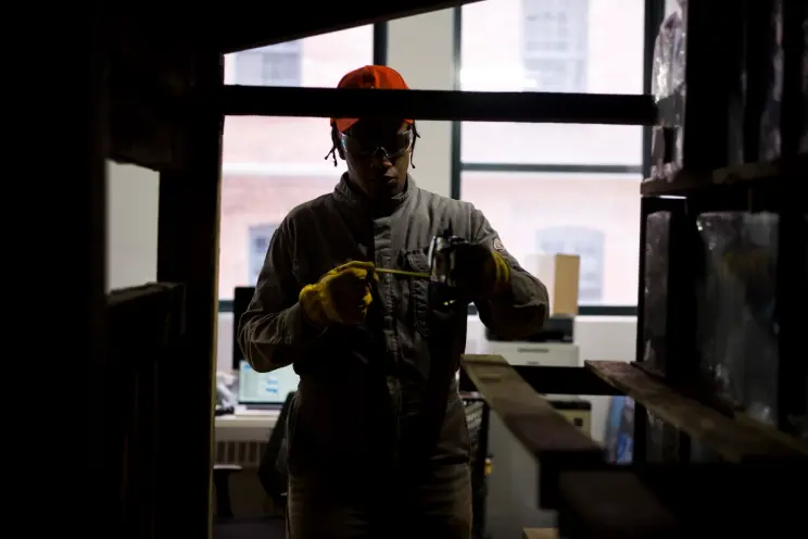 A worker in a protective suit and gloves measures something with a tool, in a dimly lit workshop.