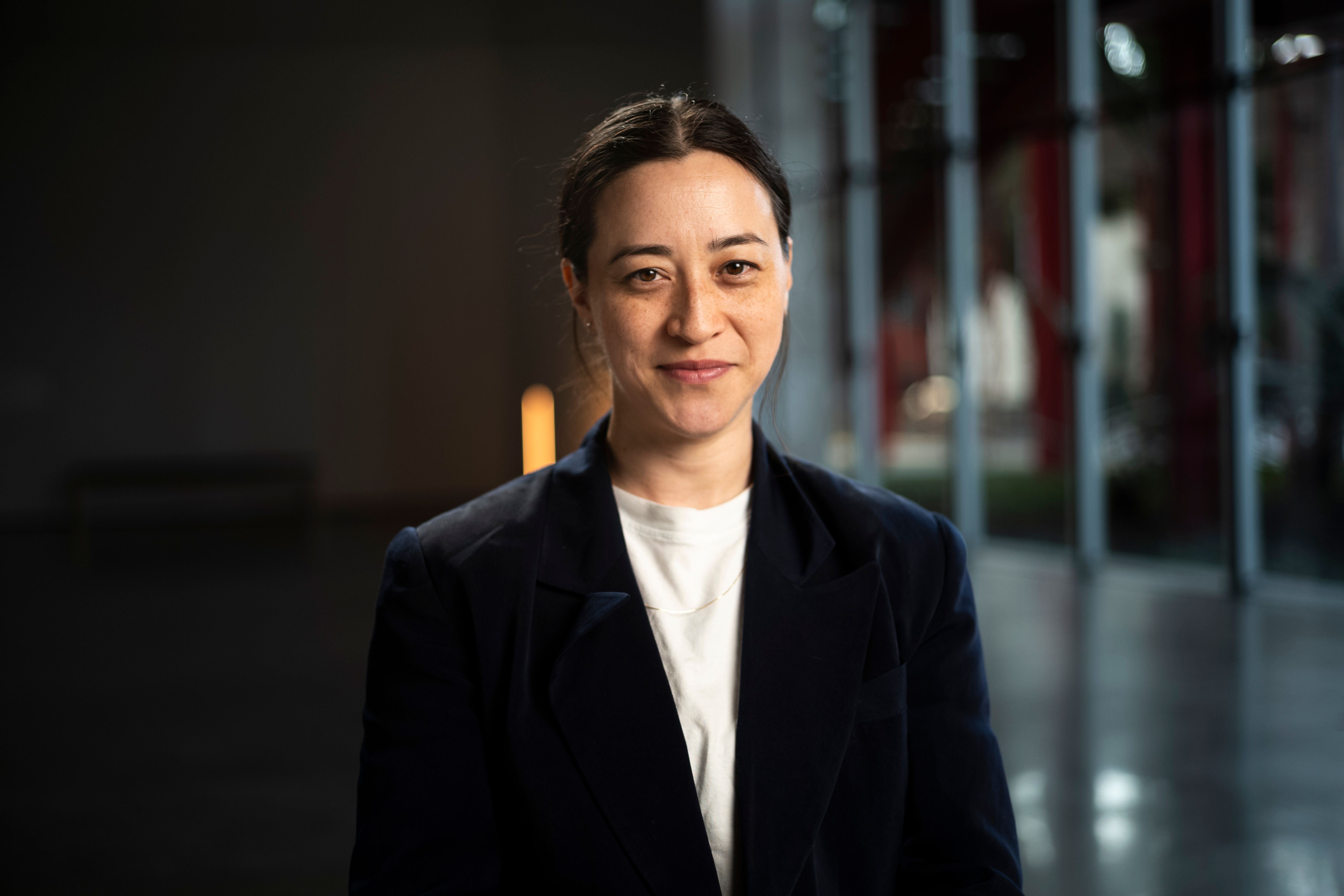 A portrait of a smiling individual wearing a dark blazer, set against a softly lit indoor background.