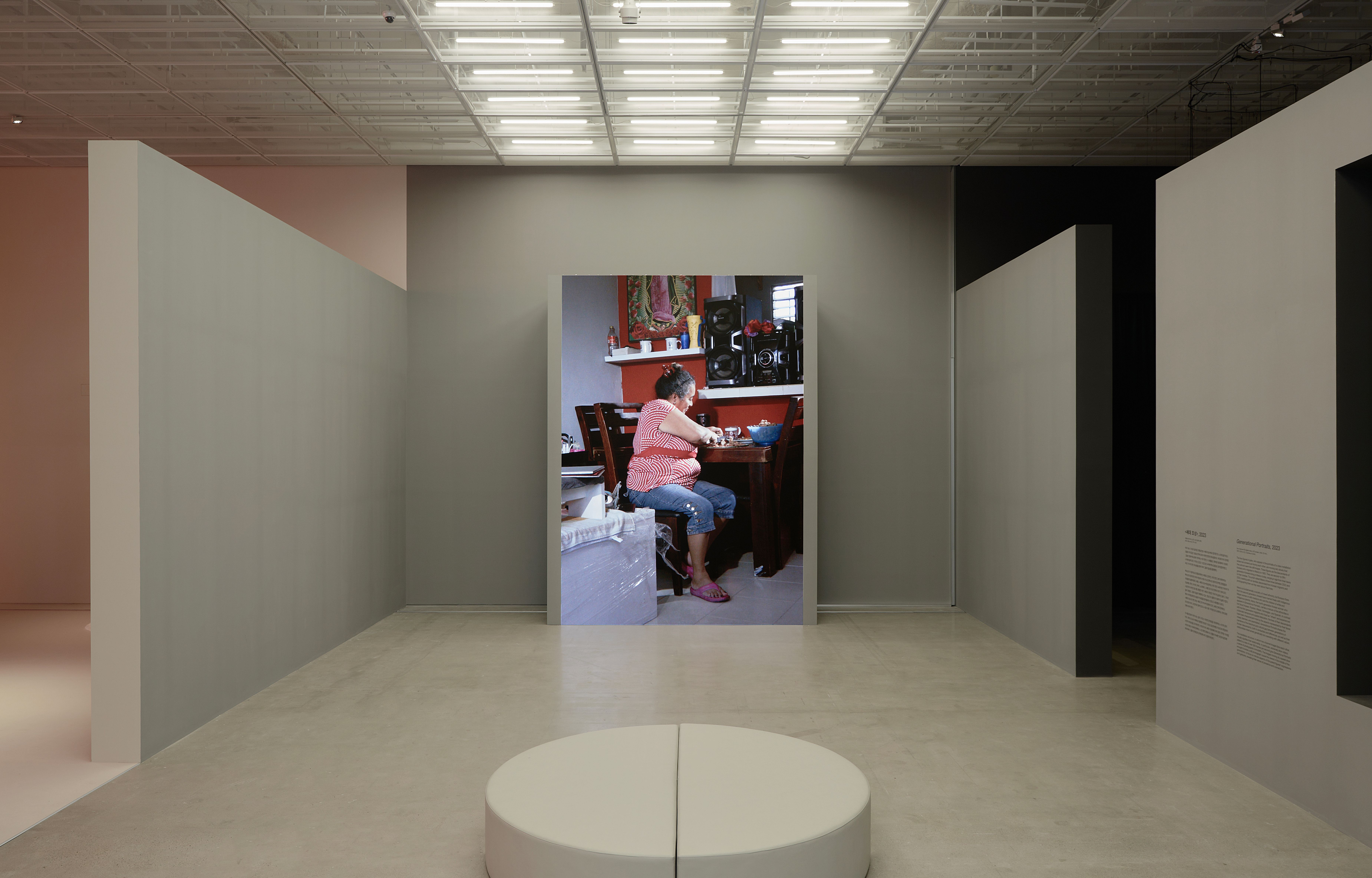 Video installation titled 'Generational Portraits': A woman sits at a wooden table in a room with a sound system above her.