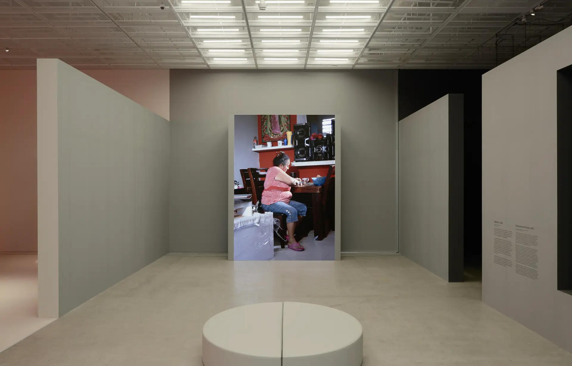 Video installation titled 'Generational Portraits': A woman sits at a wooden table in a room with a sound system above her.
