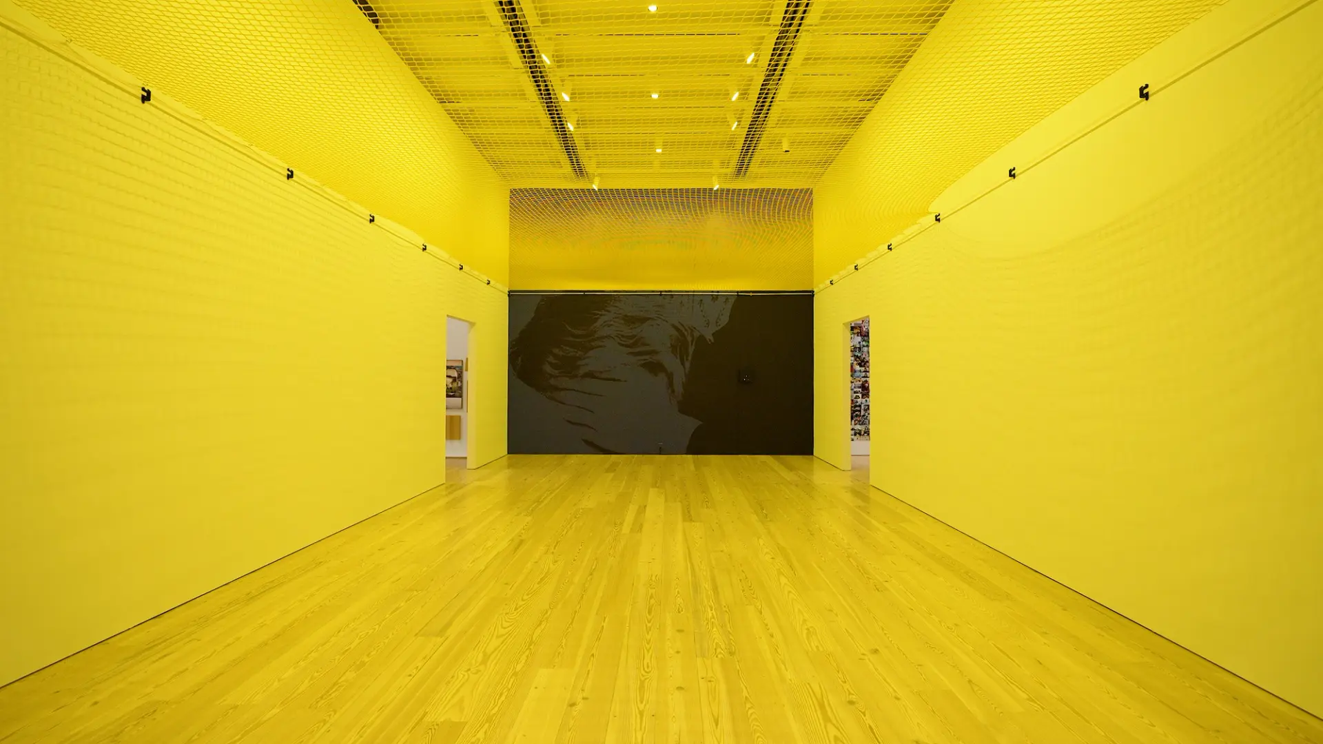 A bright yellow gallery space with a large monochrome portrait on the far wall.
