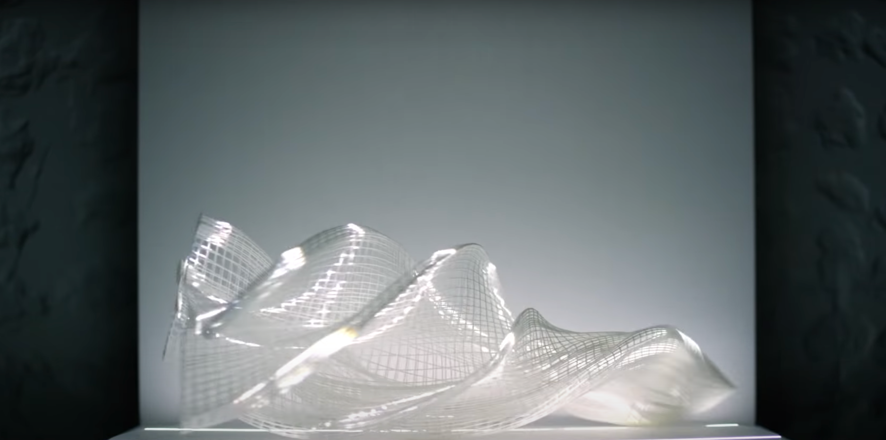 A white, net-like 3D printed sculpture on a light grey panel, surrounded by dark grey walls.