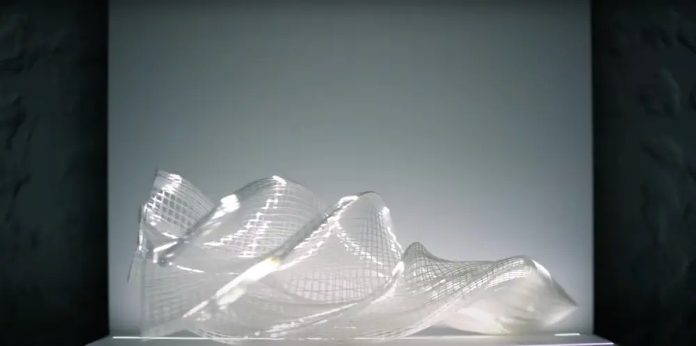 A white, net-like 3D printed sculpture on a light grey panel, surrounded by dark grey walls.