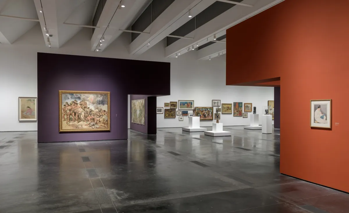 A gallery room with art pieces displayed on white cubicles and mounted on orange and purple exhibition walls.