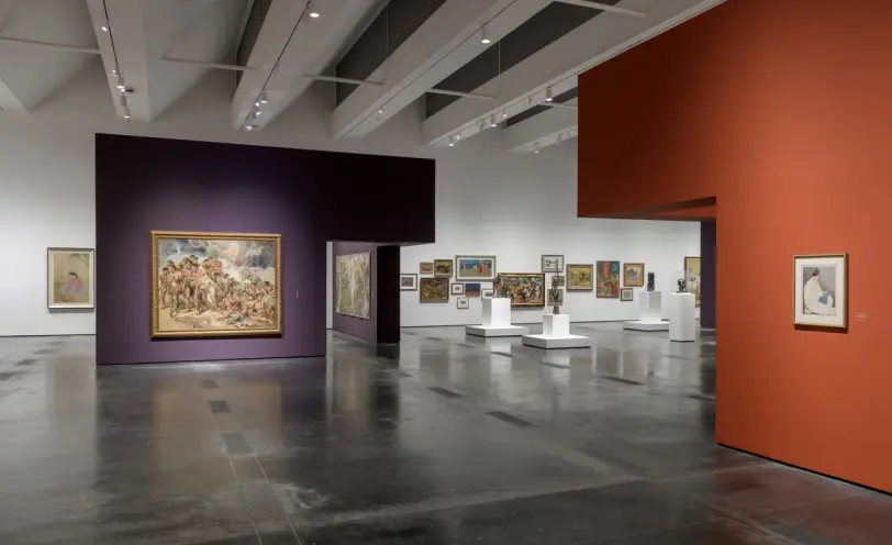 A gallery room with art pieces displayed on white cubicles and mounted on orange and purple exhibition walls.
