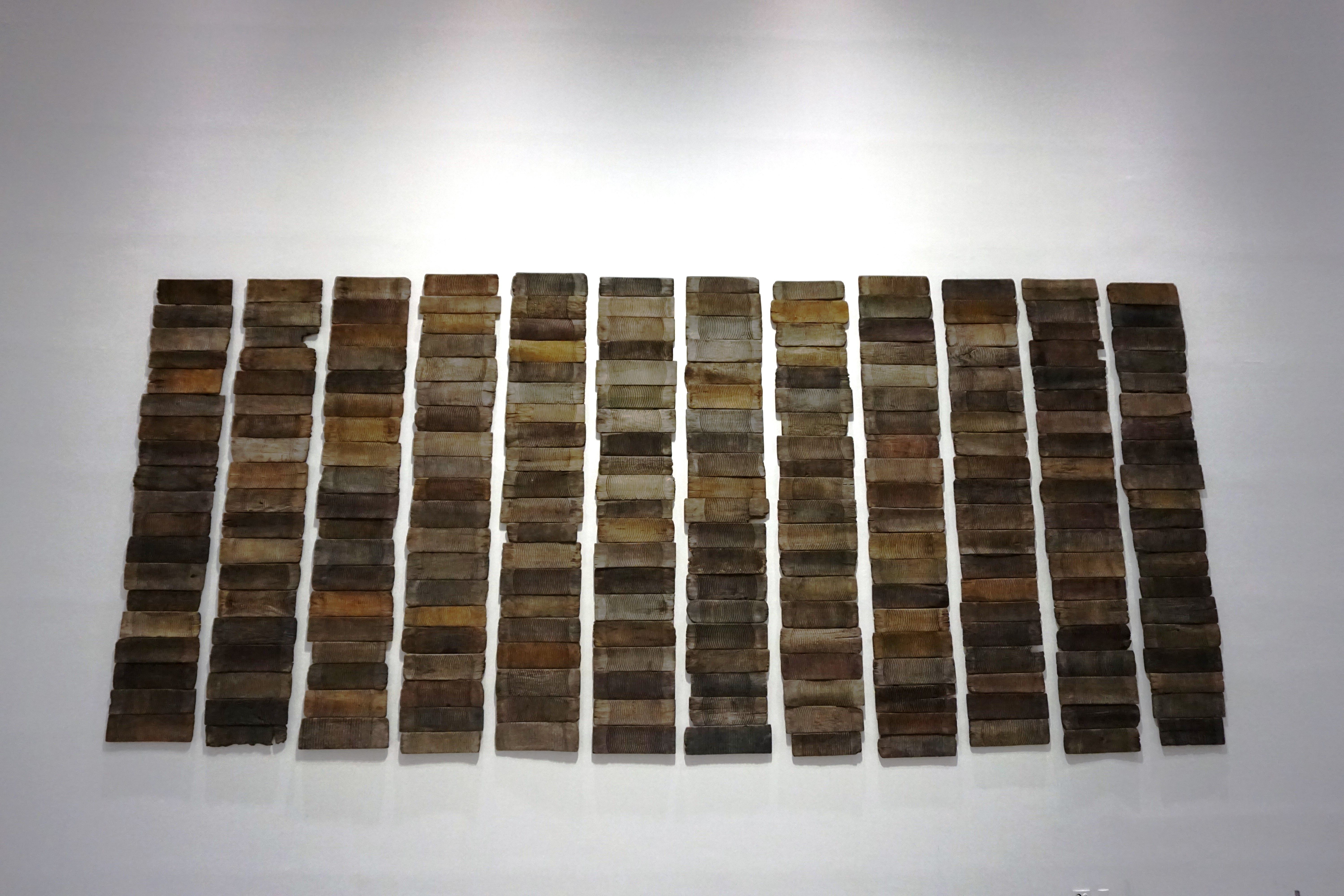 Several panels resembling wood are arranged in a grid on a pristine white wall, giving the appearance of a large-scale art...