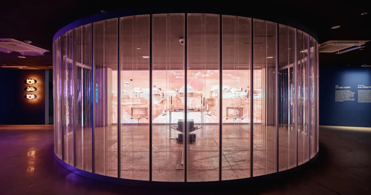 A large 3D performative apparatus-environment named "Crypton, Tour d'Epicure" stands in the center of a round room with glass walls. The structure, which includes a metal pedestal and crystal balls, is placed near a desk facing a big screen.