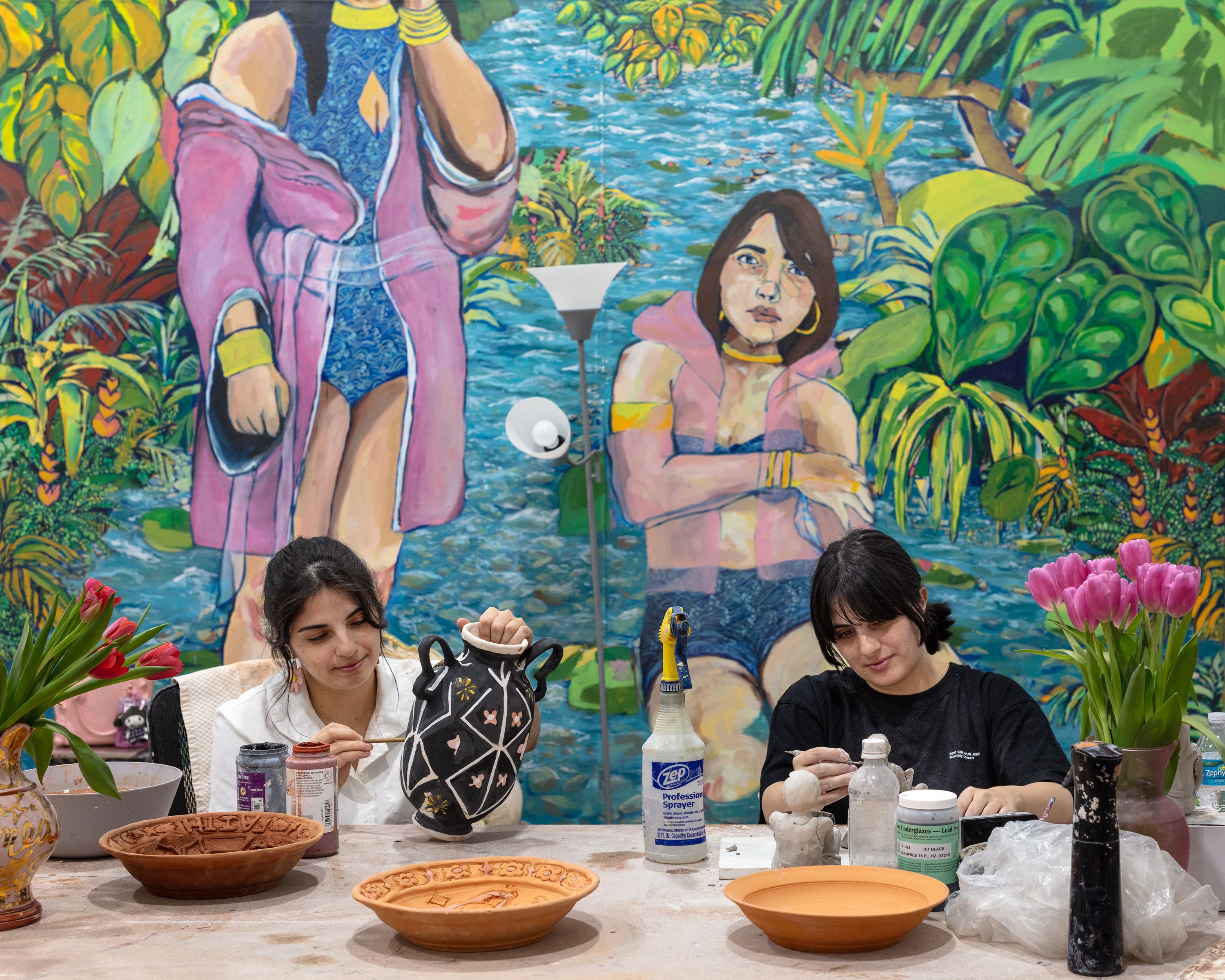 Two people sit at a table glazing ceramic vessels.