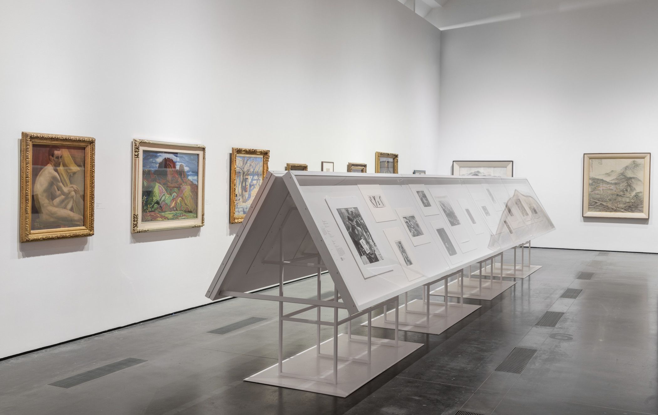 A gallery room displaying art pieces within white frames mounted on metallic stands, a part of the "The Space Between: The...