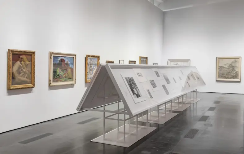 A gallery room displaying art pieces within white frames mounted on metallic stands, a part of the "The Space Between: The...