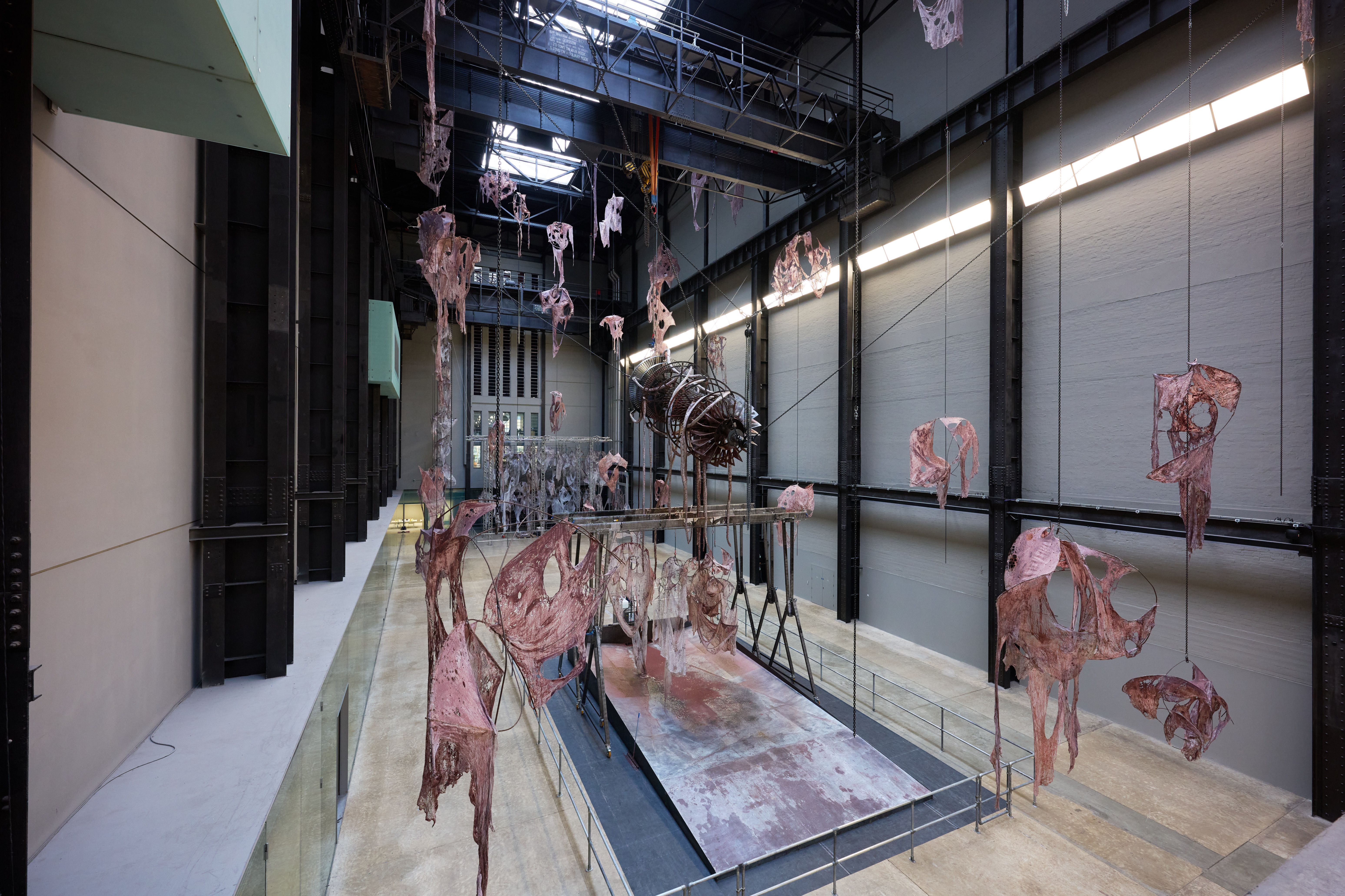 View of large installation featuring suspended fleshy, skeletal forms in a spacious industrial gallery.