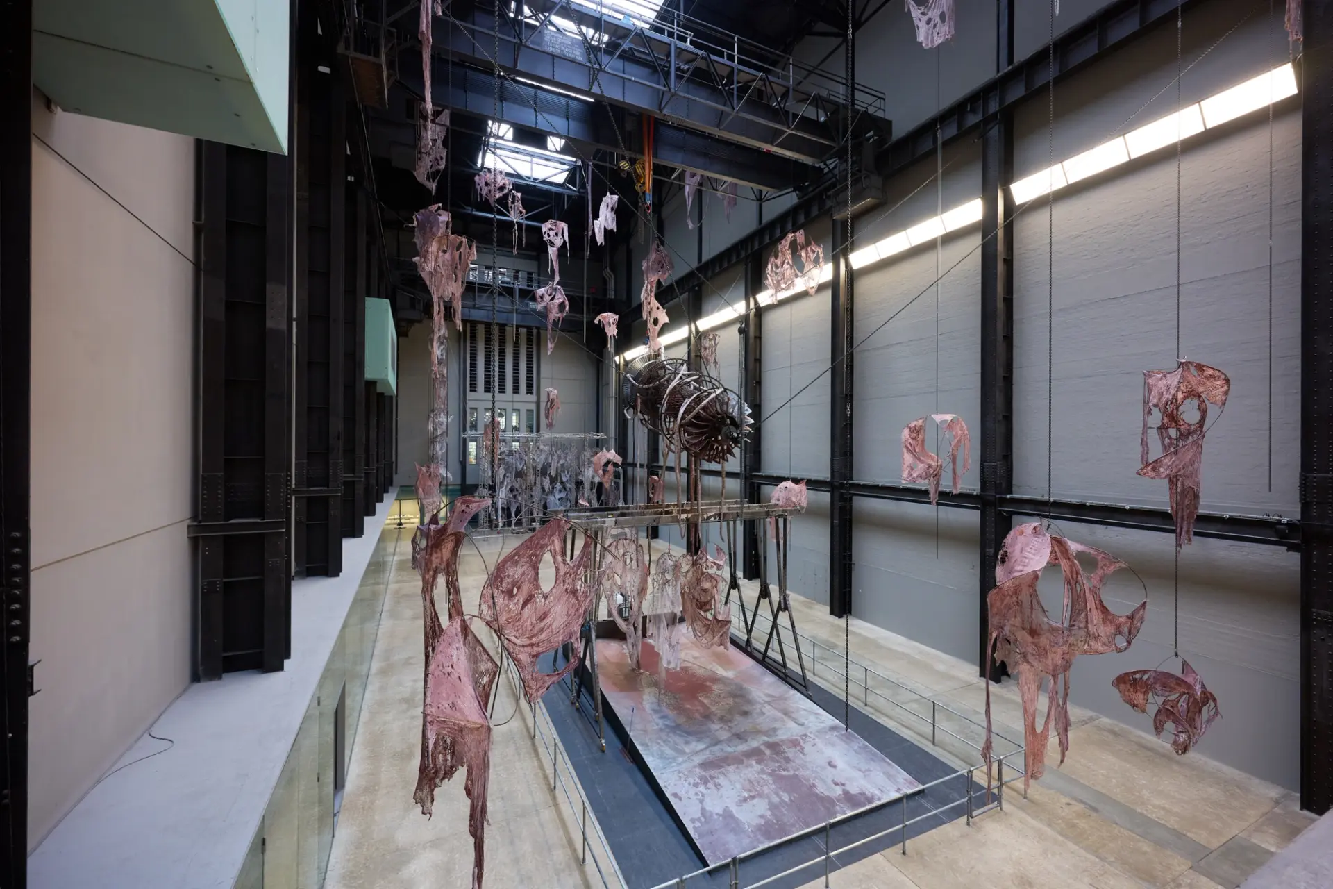 View of large installation featuring suspended fleshy, skeletal forms in a spacious industrial gallery.