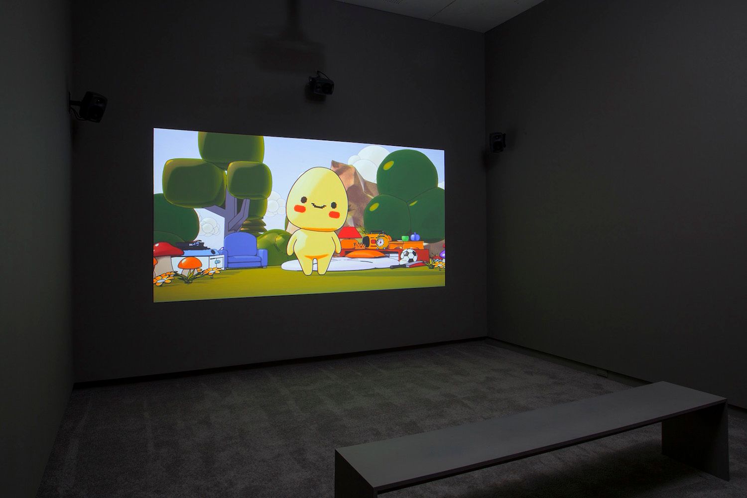 A dark room with a bench facing a large screen displaying a cute animated character in a colorful outdoor setting.