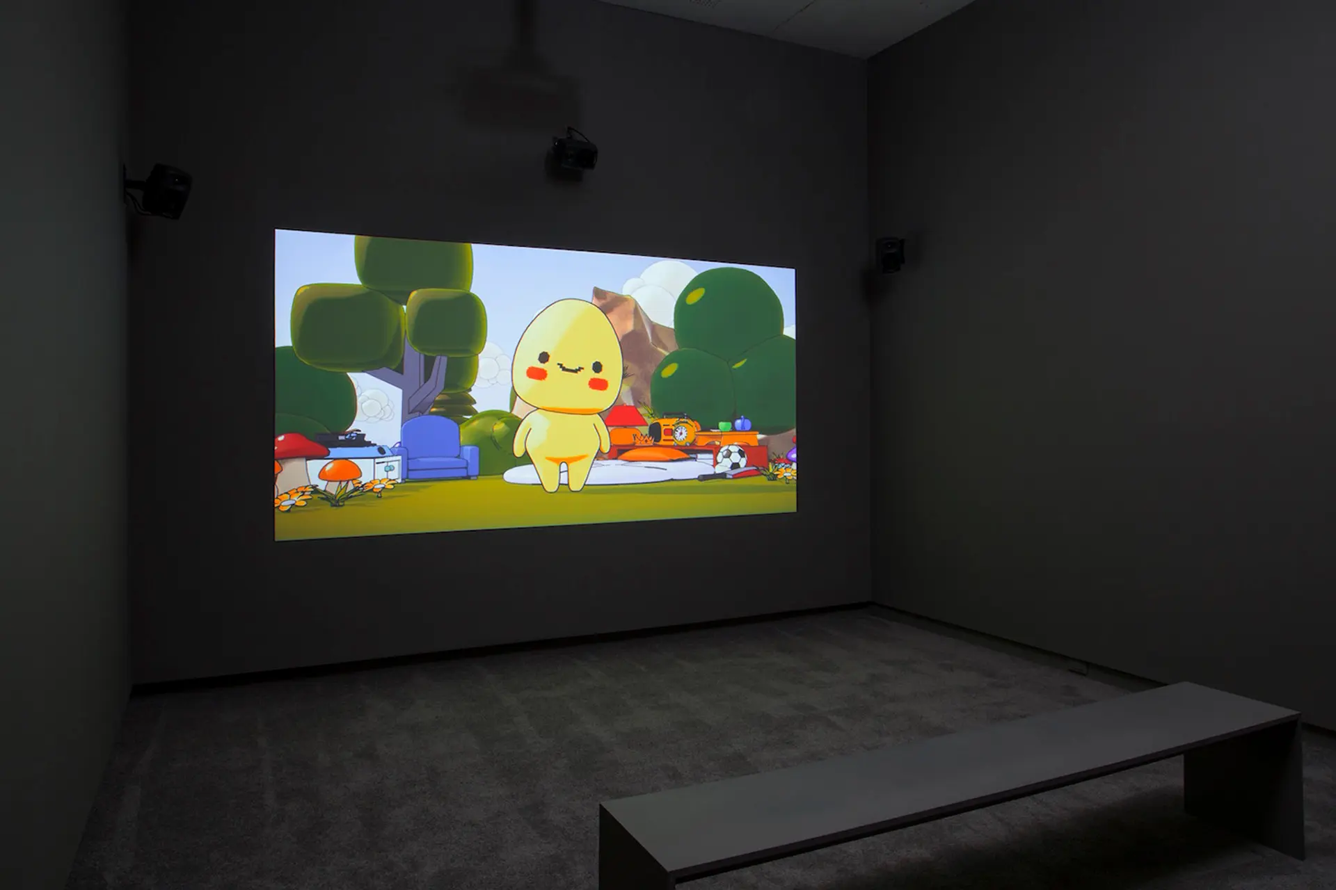 A dark room with a bench facing a large screen displaying a cute animated character in a colorful outdoor setting.