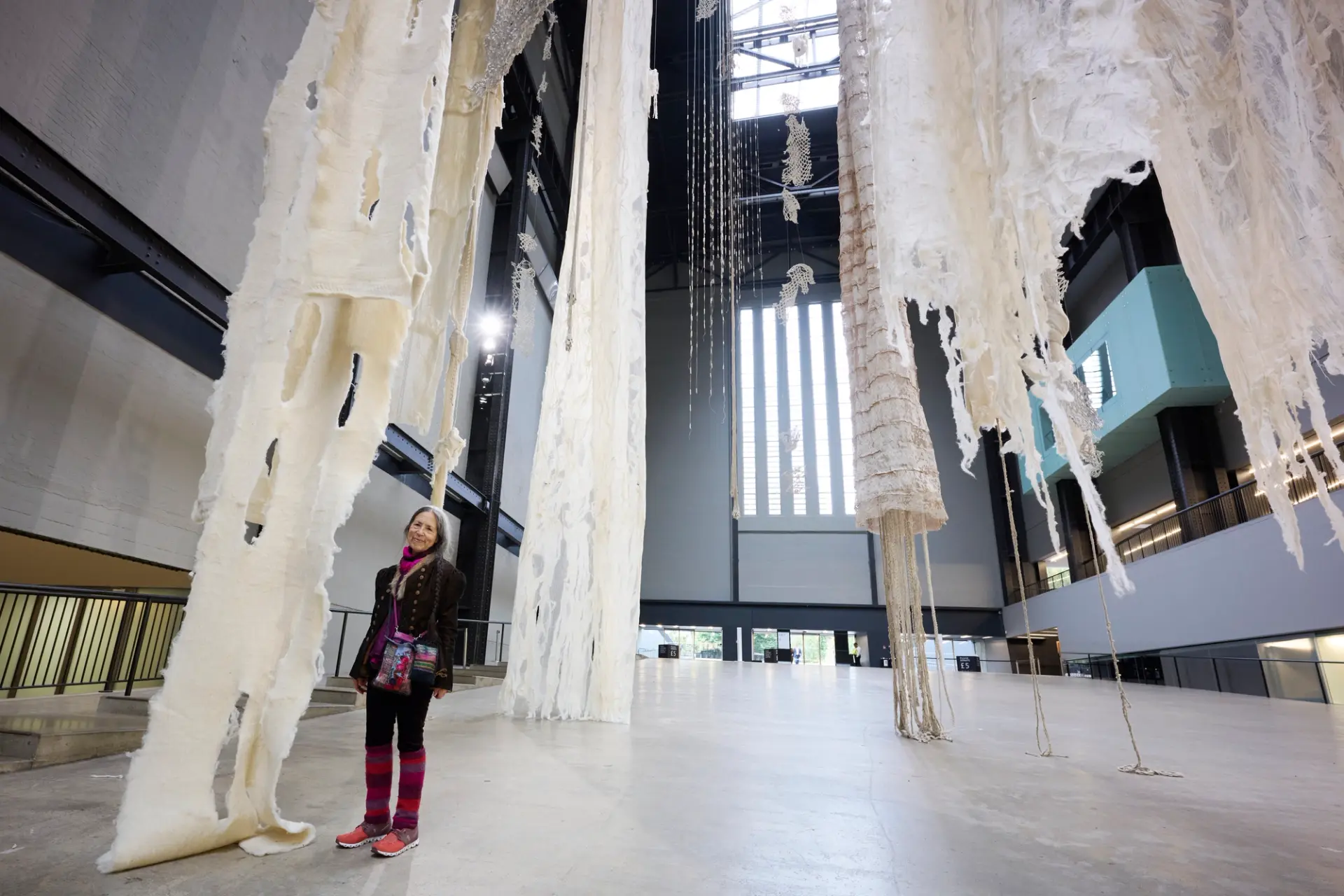 A woman stands beside her large installation of long, ripped white material which dangle from the gallery's ceiling.