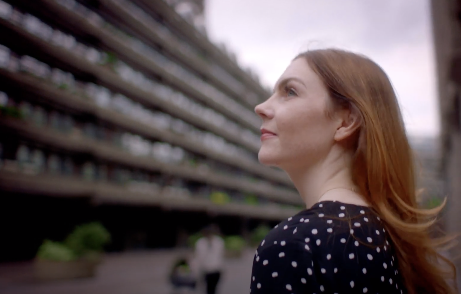 A person with red hair, captured in profile, gazes upward. In the softly blurred background, a wide seven-story building l...