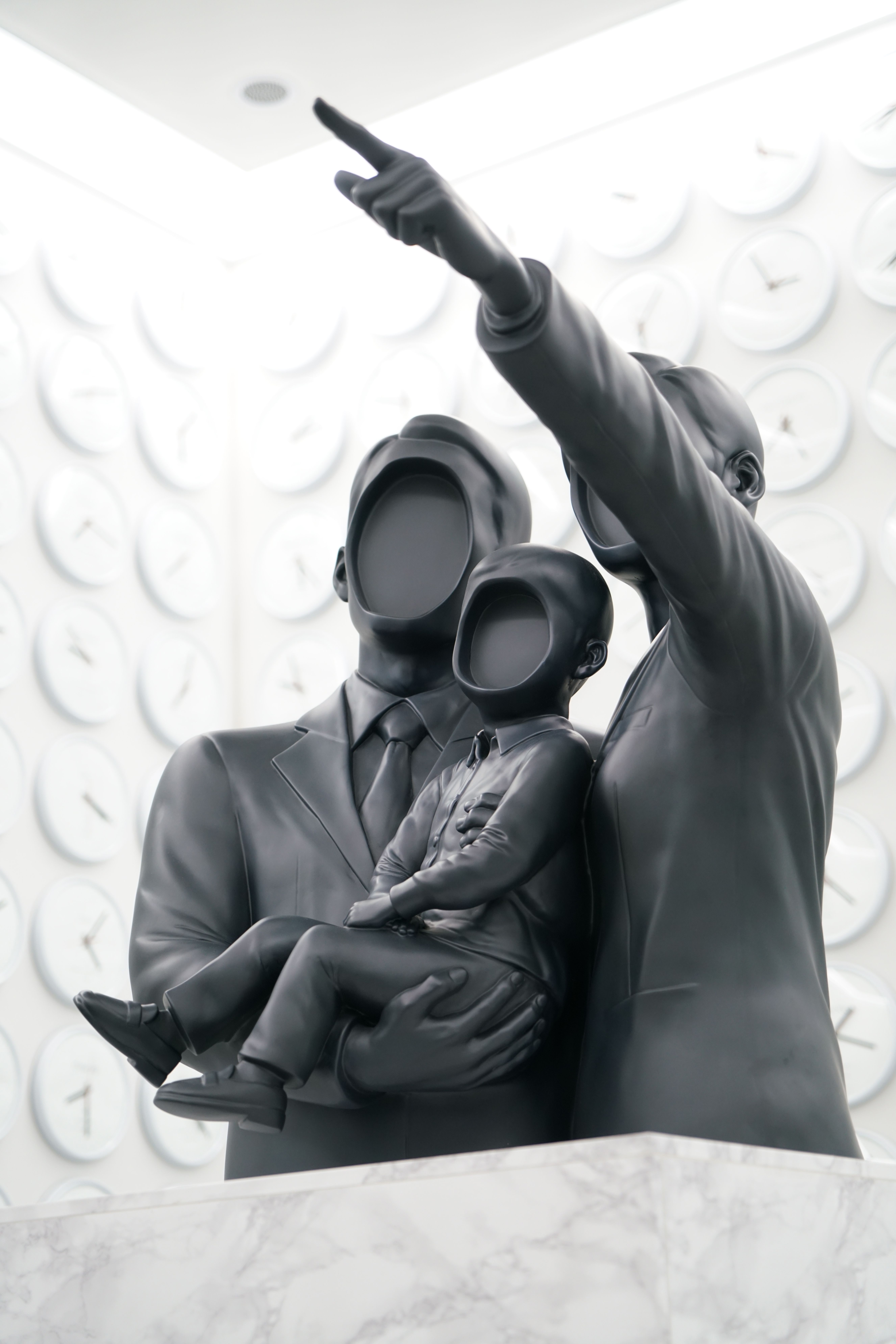 A minimalist black sculpture depicting a faceless family with two adults and a child stands against a white wall filled wi...