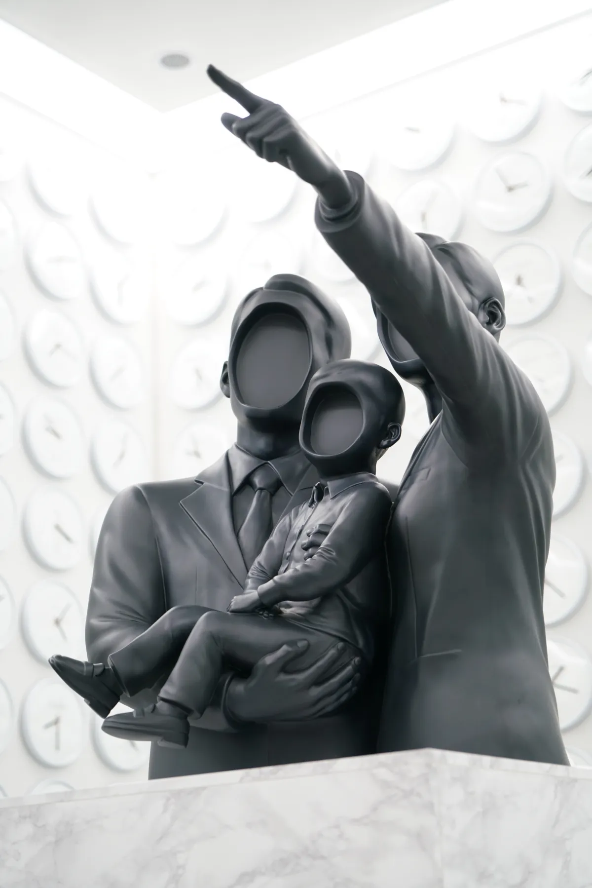 A minimalist black sculpture depicting a faceless family with two adults and a child stands against a white wall filled with clocks showing various time zones.