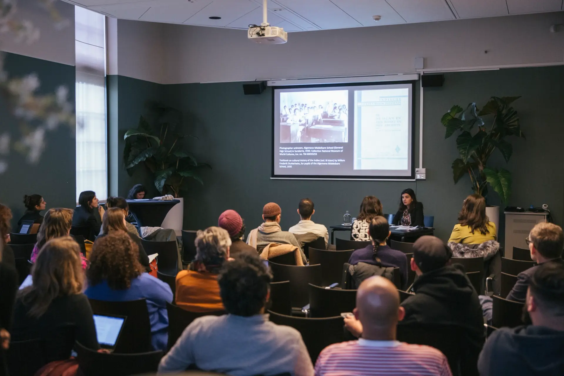 A person seated at a desk addressing a seated audience in a room with dark-green walls, a multimedia projector displaying a presentation.