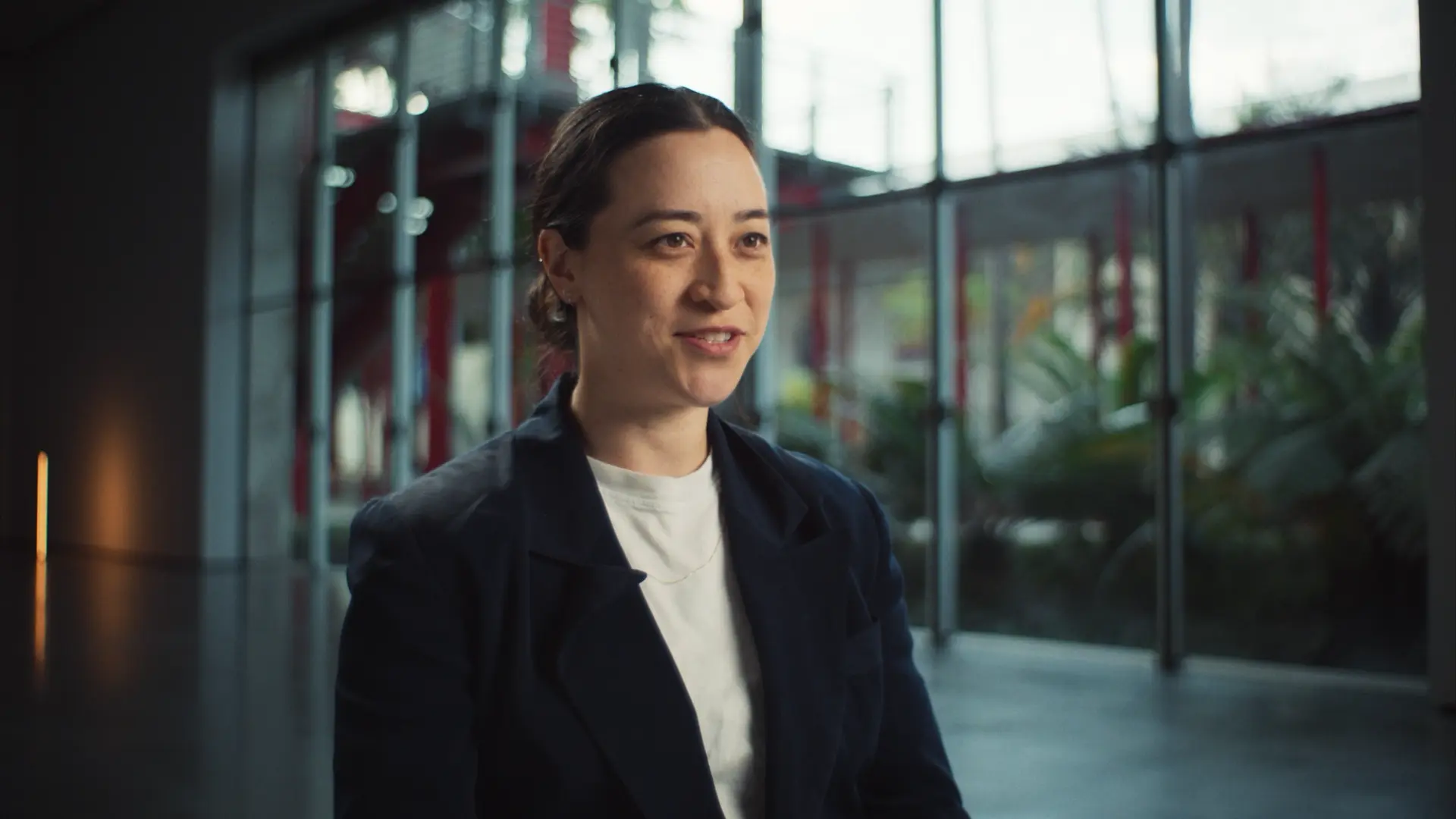 Person speaking while seated in a modern, well-lit space with large windows and greenery in the background.