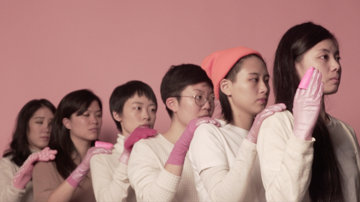 A line of individuals wearing pink gloves, each resting their hand on the shoulder of the person in front of them, standin...