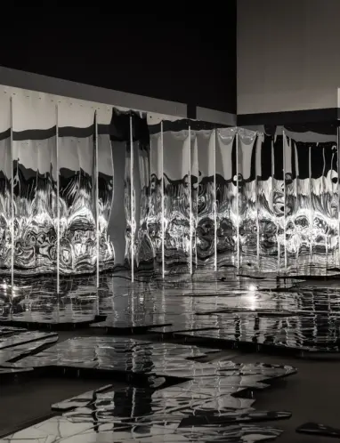 A curvilinear structure composed of reflective mirror walls stands in an indoor space with dark flooring. The mirror surfa...