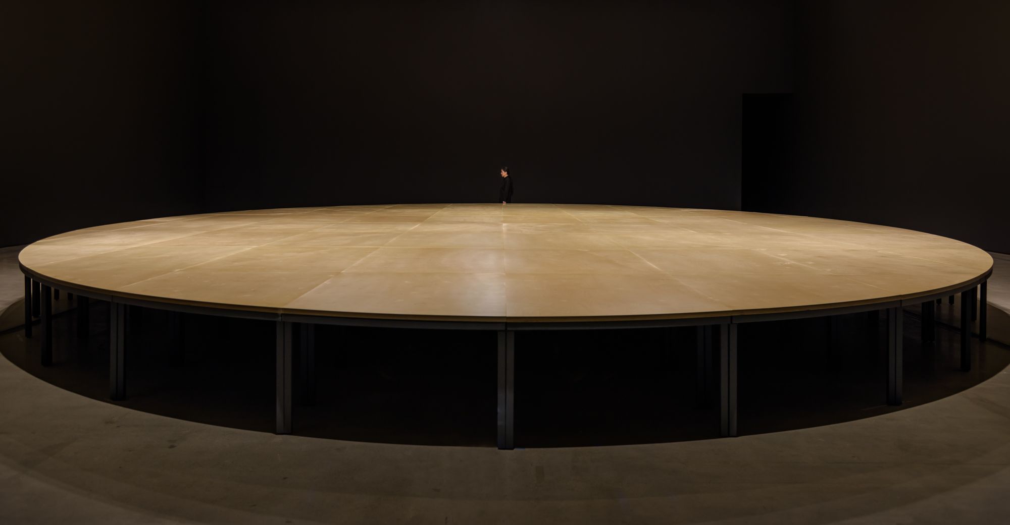 An immense, round wooden table occupies the space, highlighting the intricate grain patterns and rich brown tones of its s...
