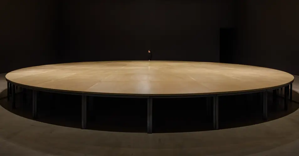 An immense, round wooden table occupies the space, highlighting the intricate grain patterns and rich brown tones of its s...