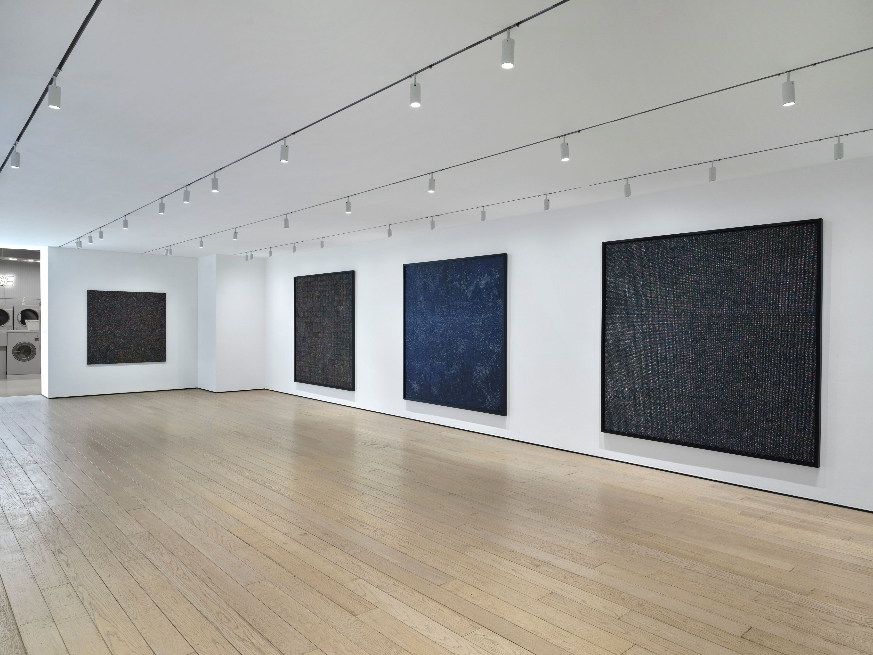 Minimal gallery with large dark abstract canvases on white walls, evenly lit, with spacious wooden floor.