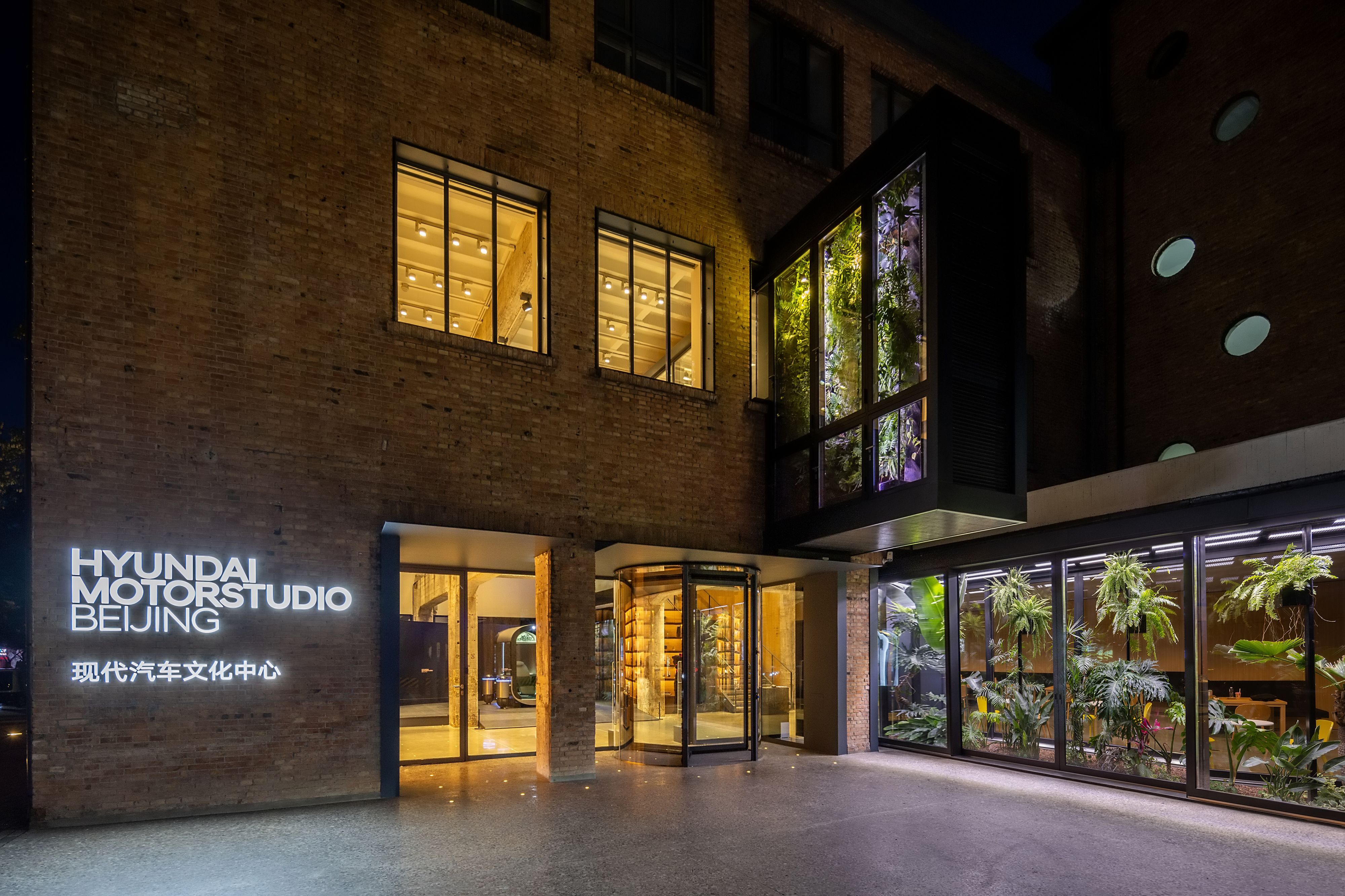 Nighttime view of the entrance to Hyundai Motorstudio Beijing, a modern cultural space housed in a brick building. Illuminated signage in English and Chinese is mounted on the left wall, while large windows reveal a warmly lit interior with wooden elements and lush greenery visible through a glass-enclosed vertical garden and a plant-filled room on the right.