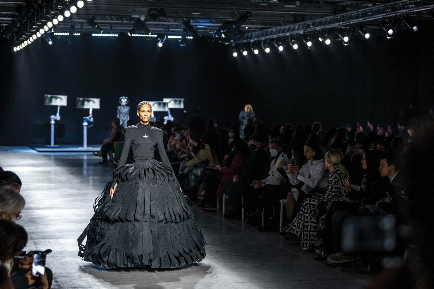 A model walks down a fashion runway in a dark dress and a high-tech bodysuit, with screens displaying eyes in the background.