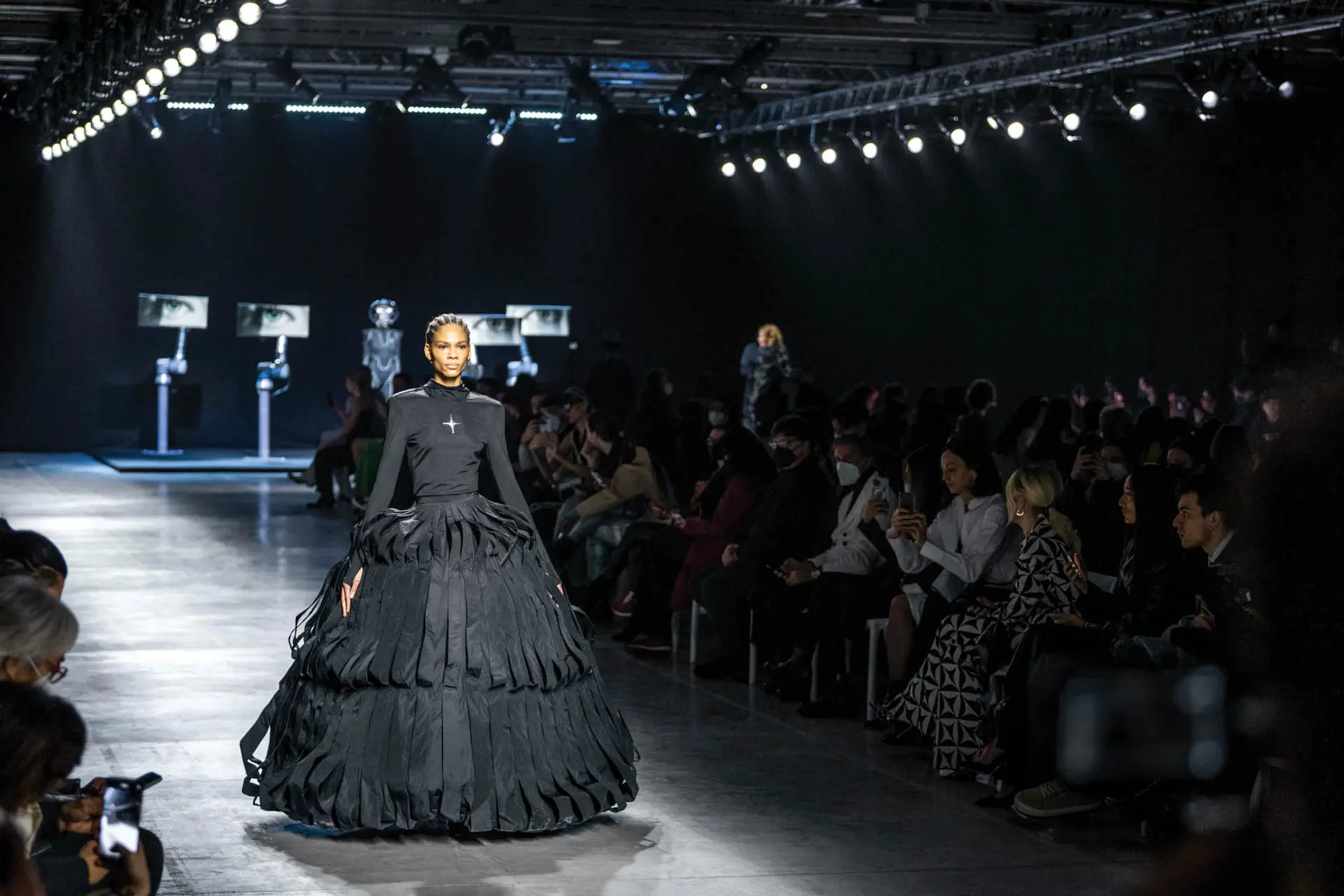 A model walks down a fashion runway in a dark dress and a high-tech bodysuit, with screens displaying eyes in the background.