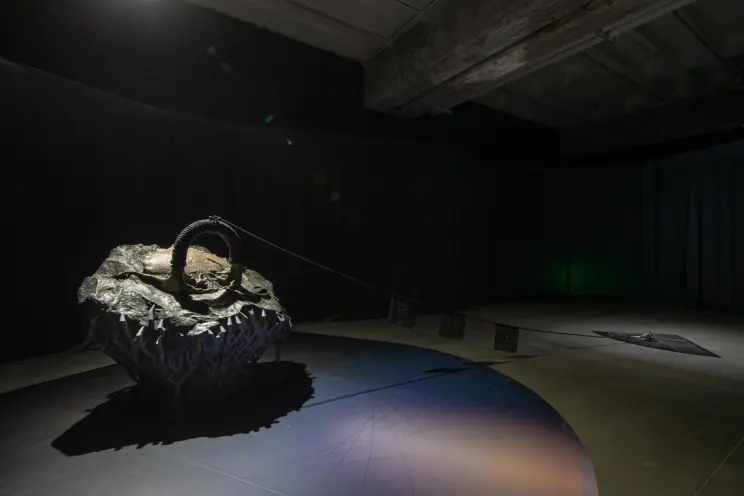 An elliptical rock tethered by chain to a triangular cover, placed on the floor of a dimly lit room.