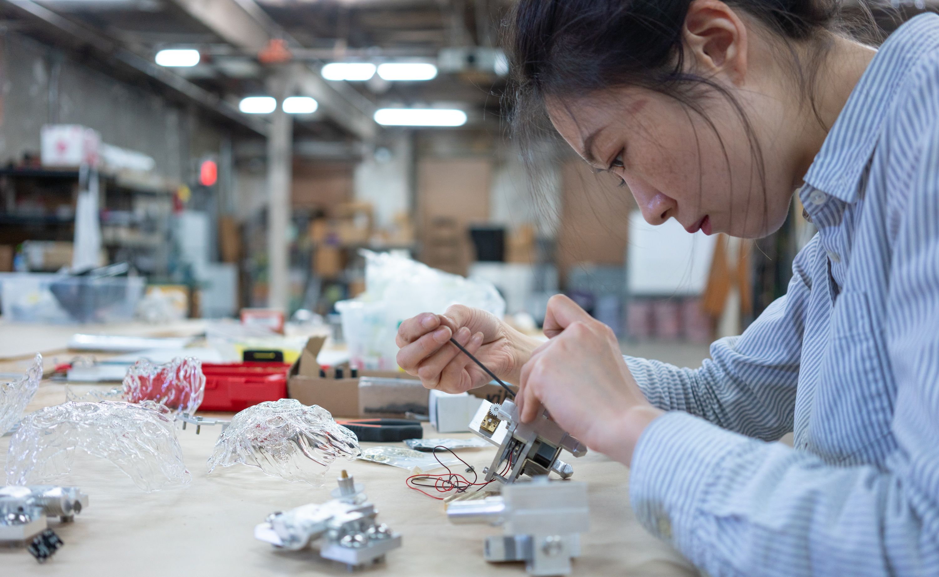 Artist working intently at a table assembling intricate metal and glass components in a workshop.