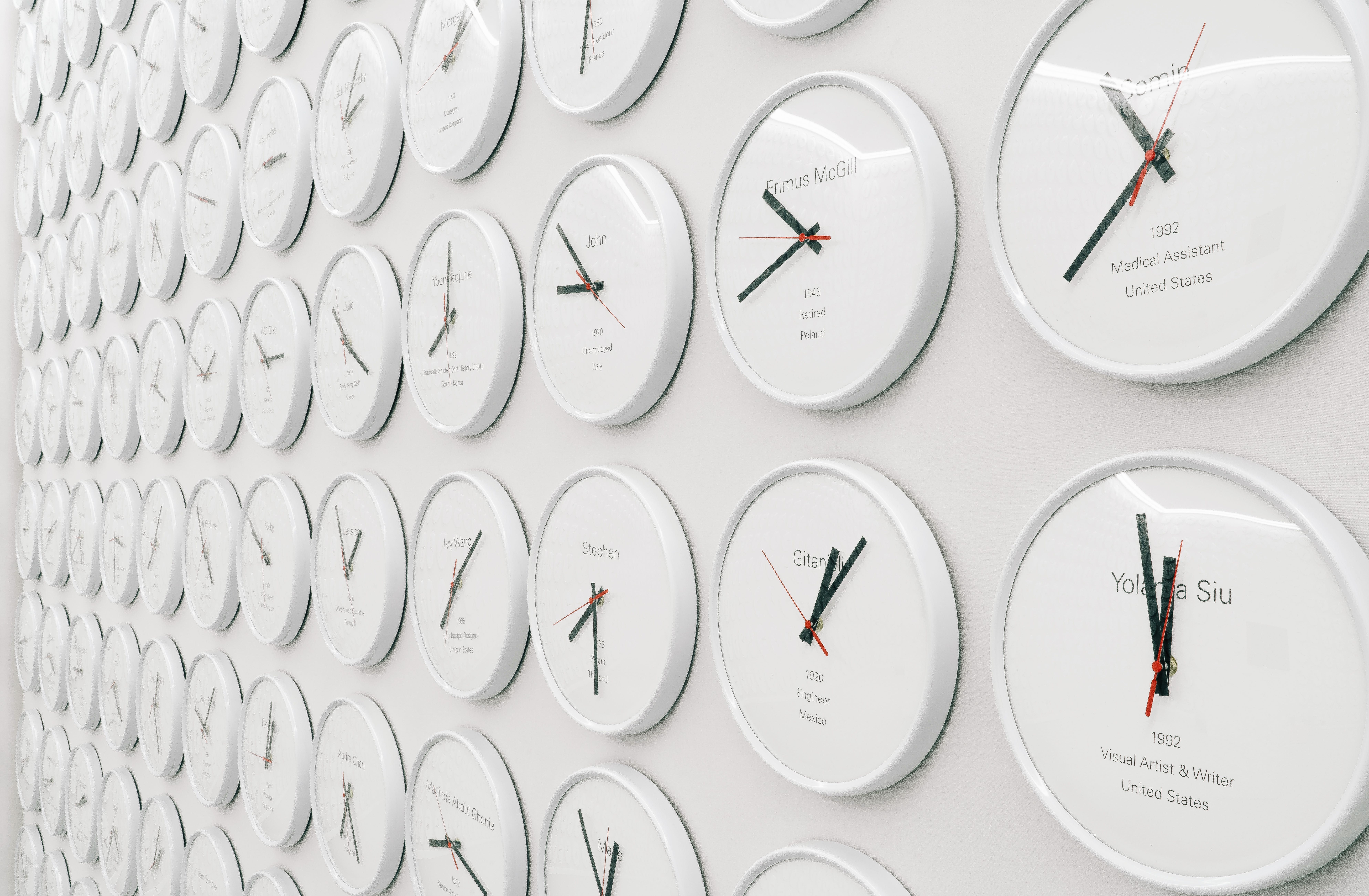 An expansive white wall is densely covered with hundreds of white-cased clocks, each showing a unique time and identified ...
