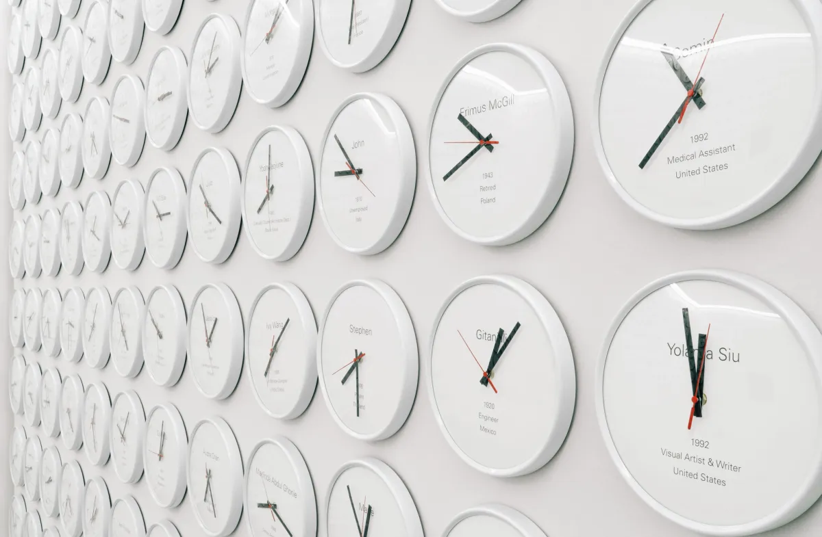An expansive white wall is densely covered with hundreds of white-cased clocks, each showing a unique time and identified by a different name. The clocks have two black hands and one red one, indicating seconds.