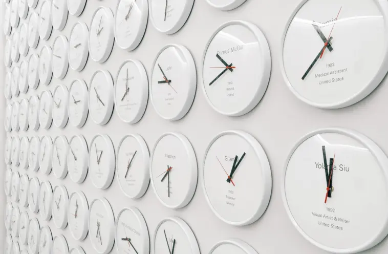 An expansive white wall is densely covered with hundreds of white-cased clocks, each showing a unique time and identified ...