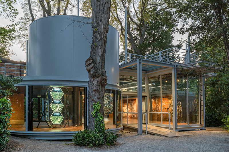 A cylindrical exhibition showroom stands outdoors, surrounded by other exhibition rooms nestled amid nature.
