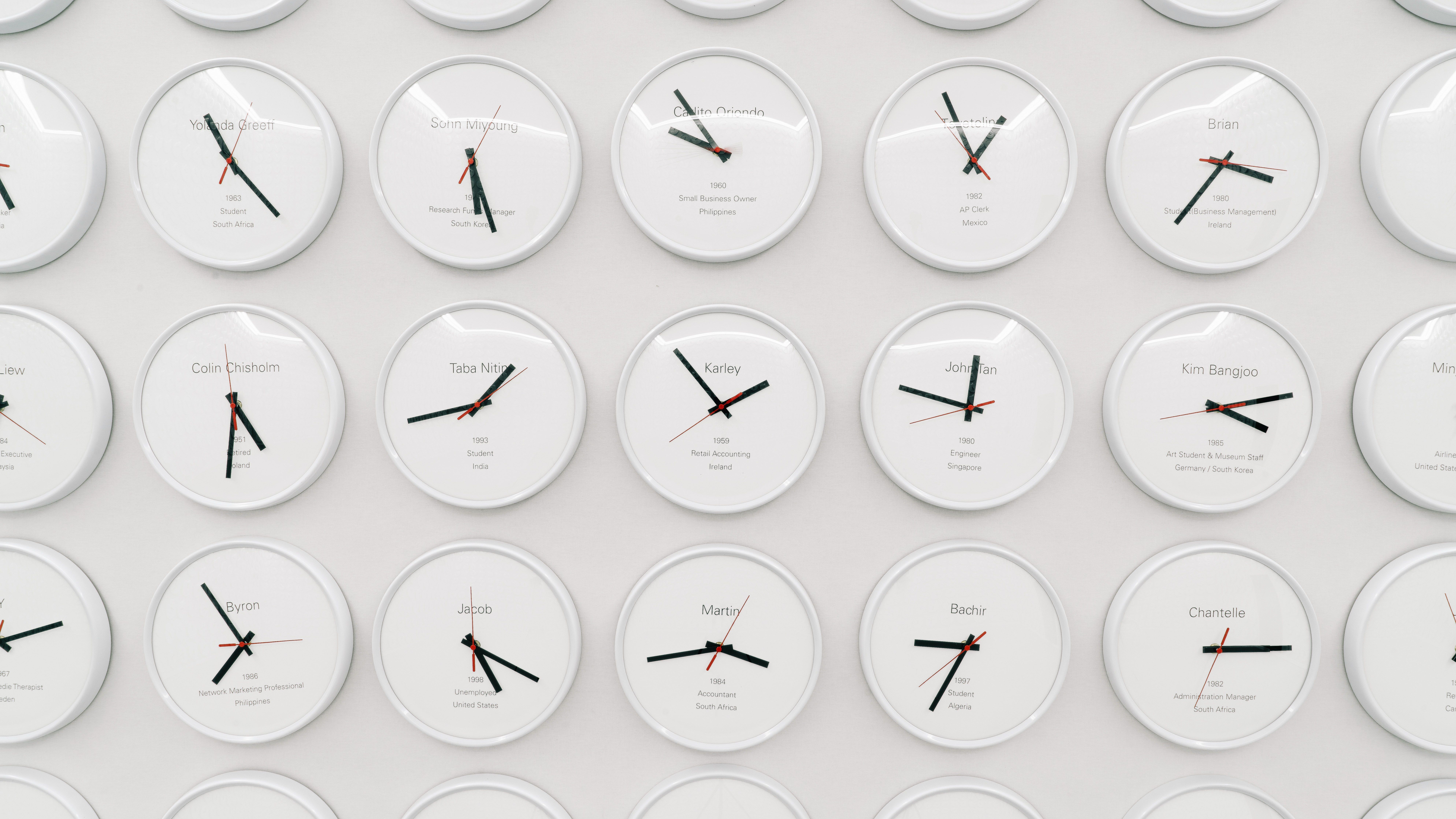 A white wall is densely populated with round clocks, each displaying a different time, indicative of various world time zo...