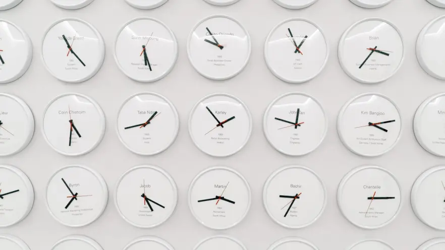 A white wall is densely populated with round clocks, each displaying a different time, indicative of various world time zo...