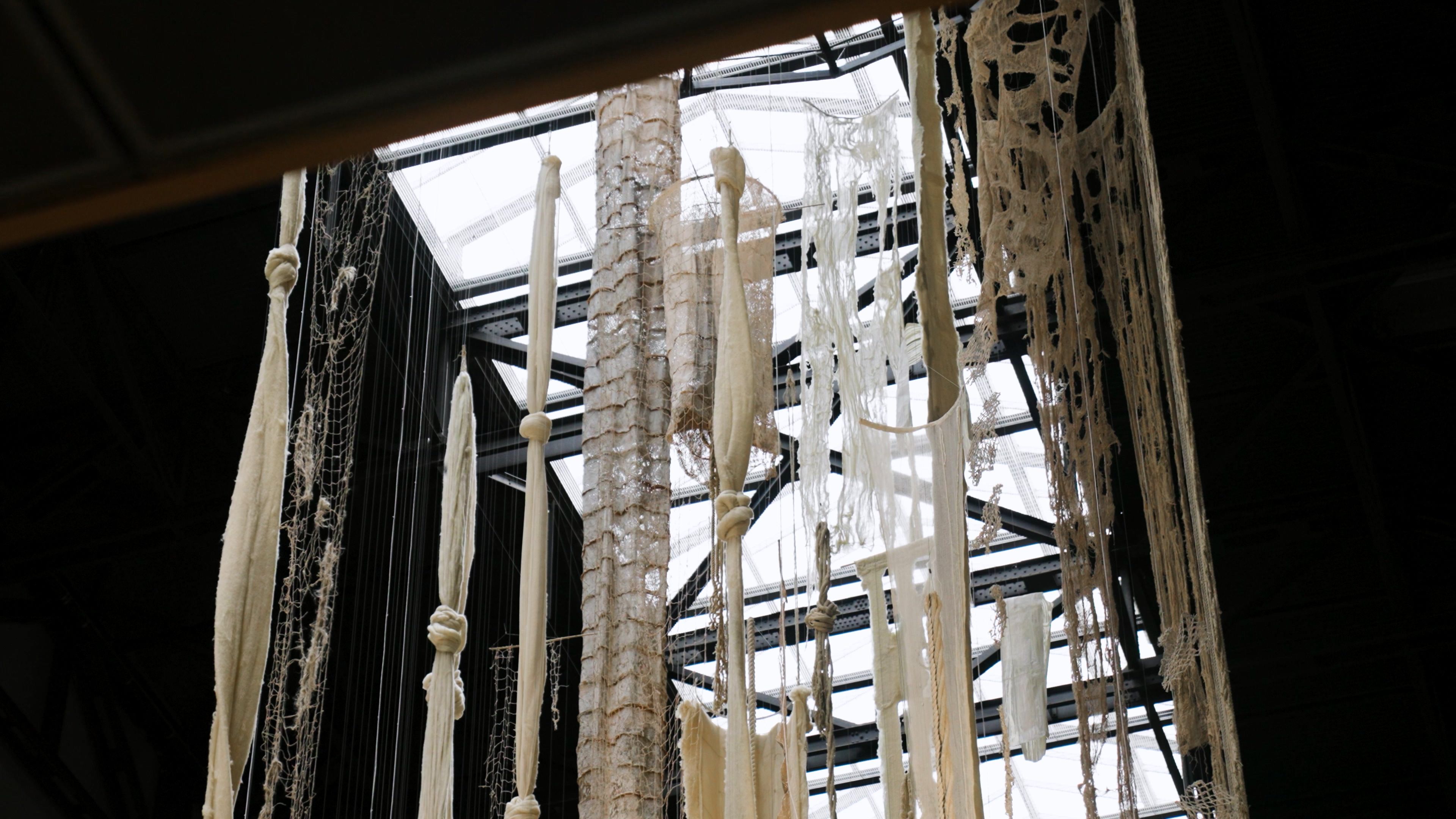 White, torn, mesh, and knotted frabics suspended from a ceiling.