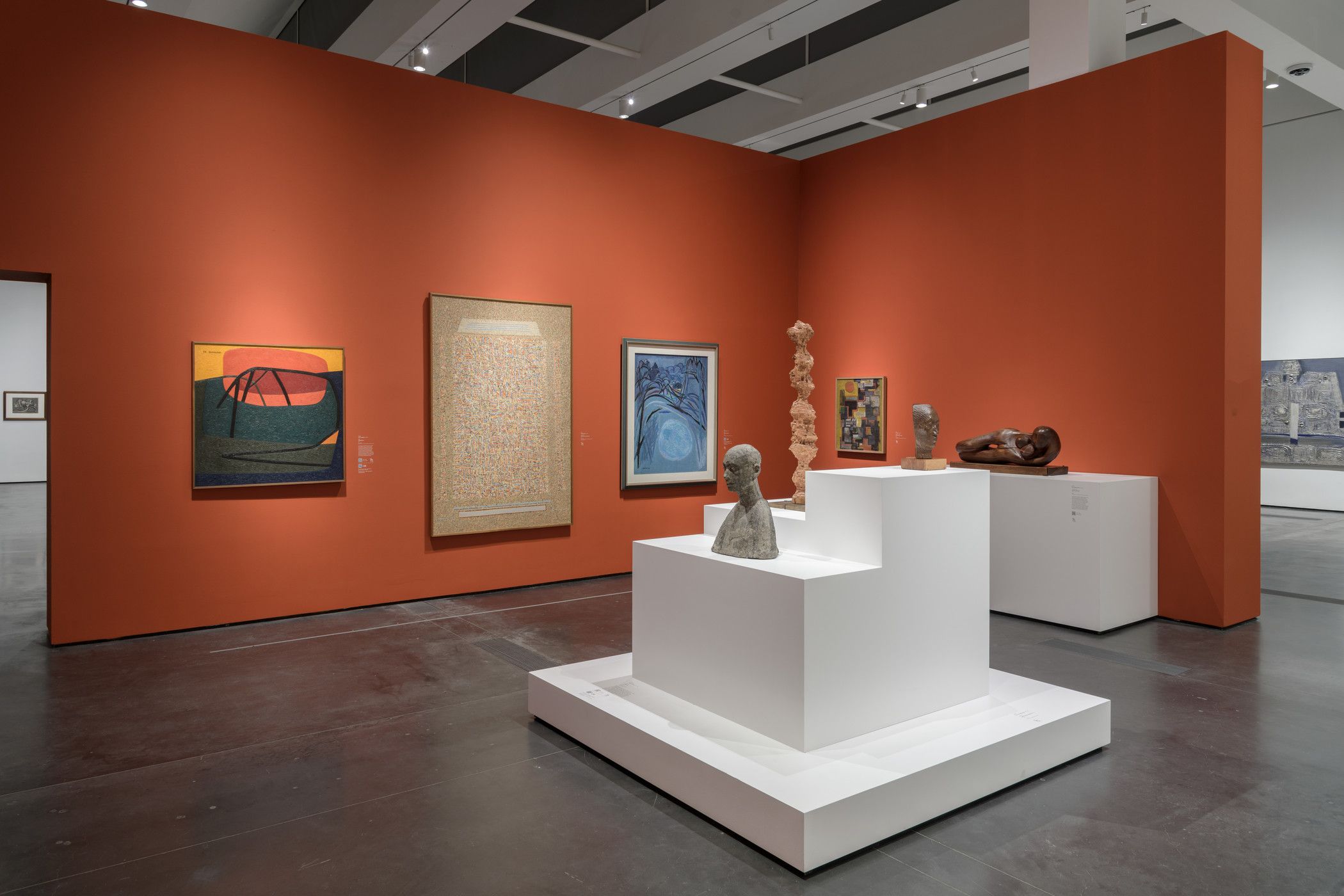 An art gallery room with various art pieces displayed against an orange exhibition wall. Additional pieces are presented o...