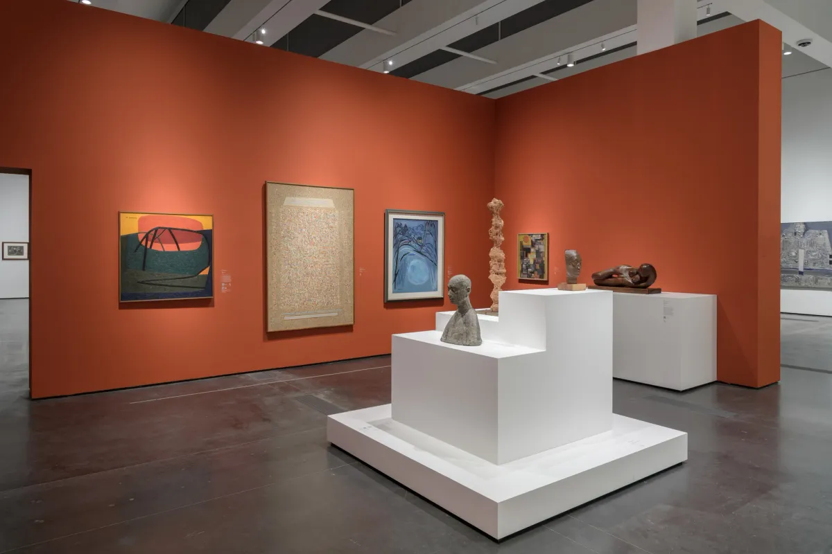An art gallery room with various art pieces displayed against an orange exhibition wall. Additional pieces are presented on white display cubes in the room's center.